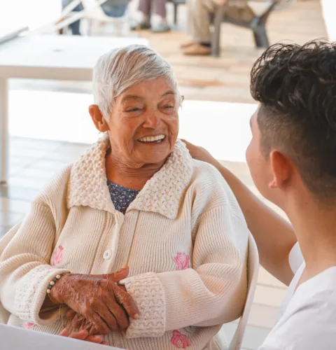 Elderly woman smiling warmly while conversing with her caregiver, sharing a moment of joy and companionship in an outdoor setting.